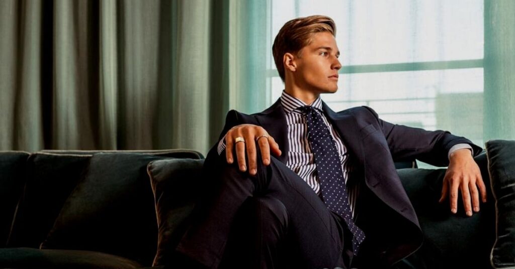 Handsome man in an elegant suit sitting relaxed on a sofa with fashion accessories like rings and bracelets, exuding modern elegance and a luxurious lifestyle.